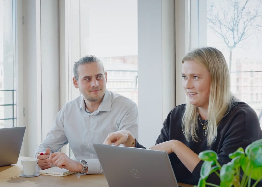 Mann und Frau mit Laptops sitzen in einem hellen Meeting-Raum und unterhalten sich miteinander.
