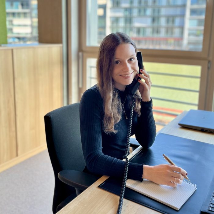 Frau am Schreibtisch in einem Büro, die einen Telefonhörer in der Hand hält und in die Kamera lächelt.