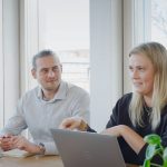 Mann und Frau mit Laptops sitzen in einem hellen Meeting-Raum und unterhalten sich miteinander.